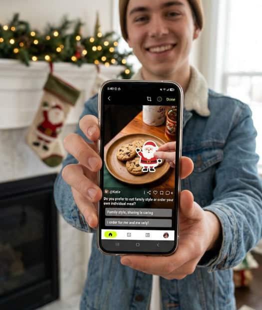 A person editing a holiday photo on their phone to add a realistic Santa sticker near a fireplace.